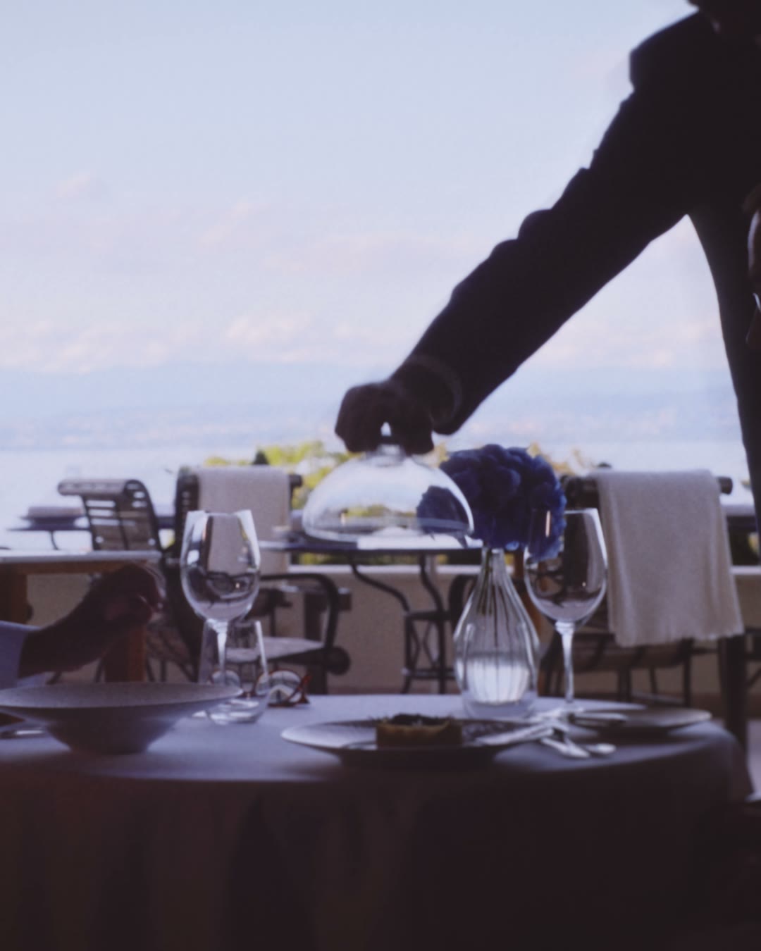 A waiter sets a plate on a table laid facing Lake Geneva